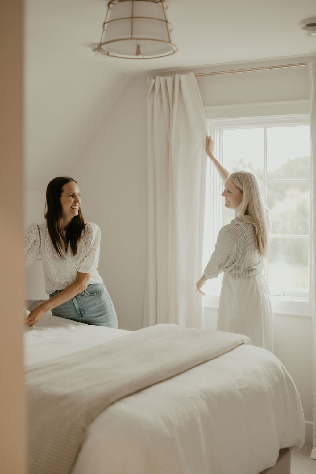 Two women in a bedroom primping the bedding and curtains